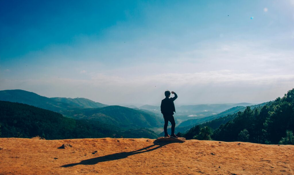 A lone traveler stands on a hilltop, gazing over a vast mountain landscape under a clear blue sky.